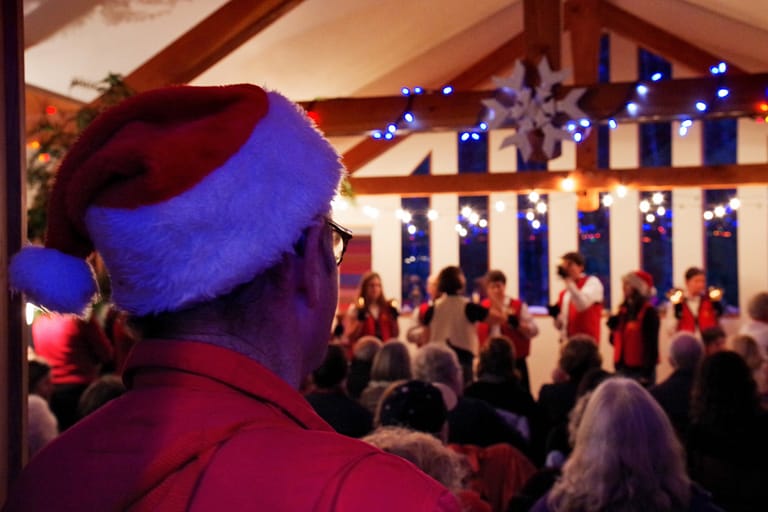 A man in a santa hat watches a handbell choir perform underneath Christmas lights.