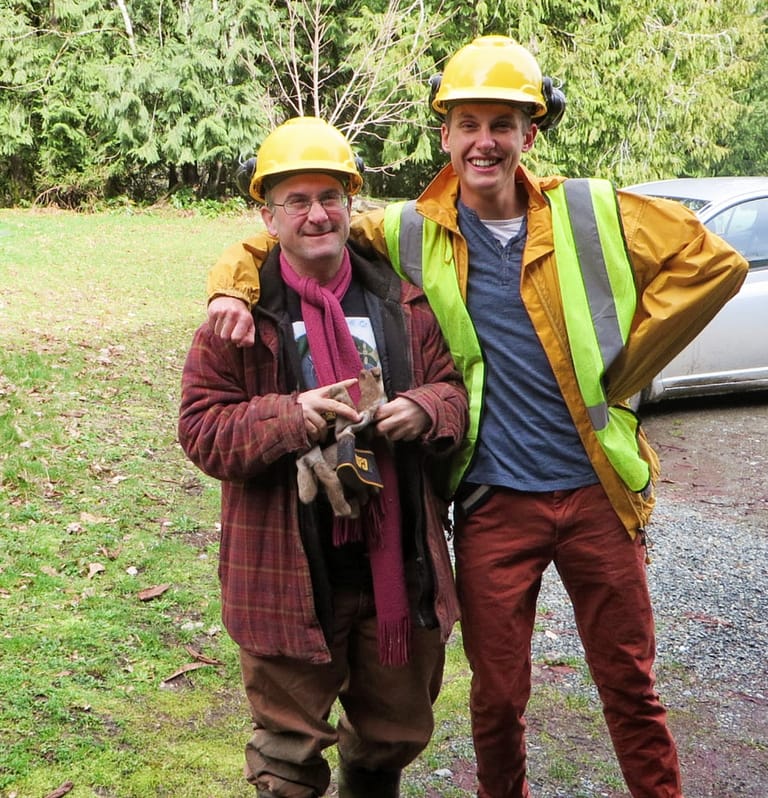 Two men stand together wearing yellow hard hats and reflector vests. They are laughing.