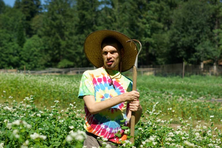 A young man stands in a field of tall grass wearing a tye dye shirt, a straw hat and holding a garden tool.